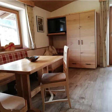 Ein gemütlicher Wohnraum mit einem Holztisch, Stühlen und einer Sitzbank. Große Fenster lassen viel Licht herein und es gibt einen Schrank mit einem Fernseher.