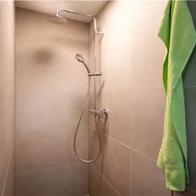 A modern shower with gray tiles and a green towel hangs next to it. The shower has a simple, clear faucet and a rain showerhead.
