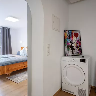 A modern room with a washing machine and an artistic picture on the wall. Through the open door, a bedroom with a wooden bed and curtains is visible.