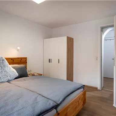 A modern bedroom with a large bed, a pillow, and a wooden wardrobe. The walls are bright and the room feels inviting and spacious.