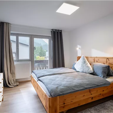 A cozy bedroom with a wooden bed and gray bedding. Large windows provide plenty of daylight and a view outside.