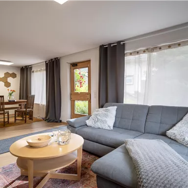 A modern living room with a cozy gray couch and a round table. Large windows let in a lot of light, and the space feels inviting.