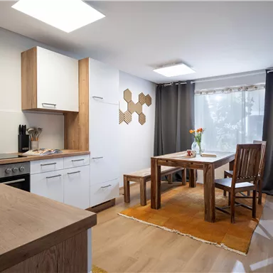 A modern kitchen with a dining area and a wooden table. Large windows provide plenty of daylight and a warm atmosphere.