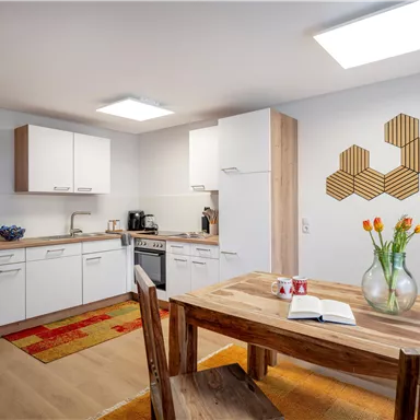 A modern kitchen with white cabinets and wood details. A wooden table with fresh flowers and a book is visible in the foreground.