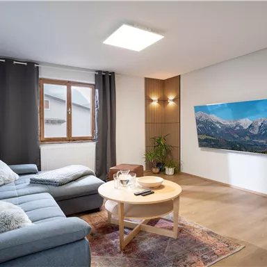 A cozy living room with a blue sofa and a round table. A television hangs on the wall, and a window lets in daylight.