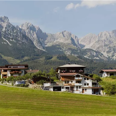 A picturesque mountain landscape with impressive peaks in the background. In the foreground, several traditional houses stand on a green meadow.
