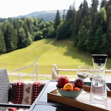 A beautiful view of a green meadow surrounded by trees. On the table, there is a carafe, a glass of water, and a bowl of fresh fruits.