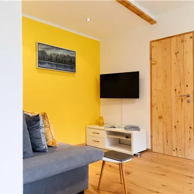 A cozy living area with a yellow wall and a modern television. There is a sofa, a small table, and a wooden door.