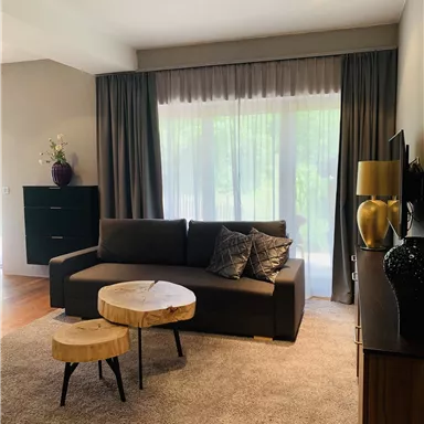Ein modernes Wohnzimmer mit einem gemütlichen Sofa und einem Couchtisch aus Holz. Große Fenster sorgen für viel Tageslicht und eine einladende Atmosphäre.