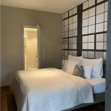 Ein modernes Schlafzimmer mit einem großen Bett und neutralen Farben. Die Wand ist mit einem grafischen Muster gestaltet, und eine Tür führt zu einem weiteren Raum.