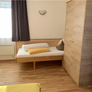 A simple bedroom with a bed and a wooden wardrobe. The room has large windows and a bright, friendly atmosphere.