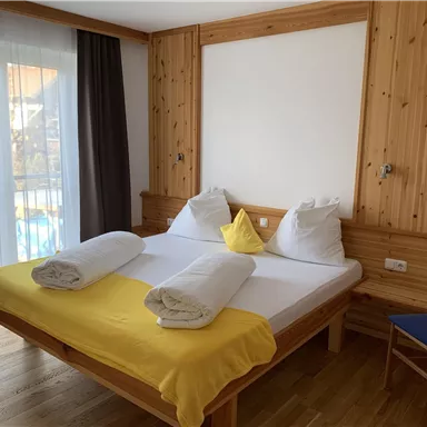 A cozy bedroom with a large bed, white pillows, and yellow blankets. Large windows let in plenty of light and provide a view outside.