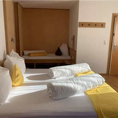 A modern bedroom with a double bed, white bedding, and yellow accents. In the background, there is a cozy seating area.