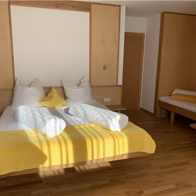 A bright bedroom with a large bed and two pillows. The walls are in warm wood tones, and there is a simple seating area.