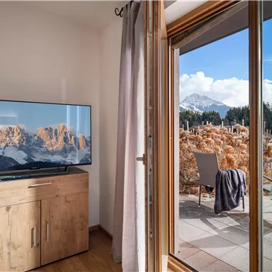 Ein gemütlicher Raum mit einem Fernseher und einem Blick auf die Berge. Draußen ist eine Terrasse mit Stühlen und einer schönen Landschaft.