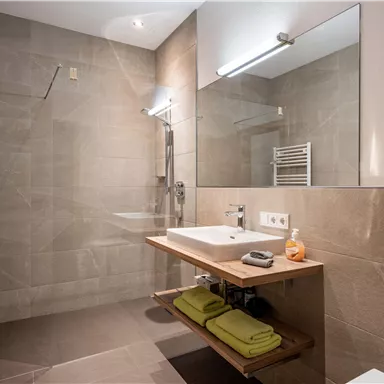 Ein modernes Badezimmer mit einer großen Dusche und einem hellen Waschbecken. Die Wände sind in neutralen Farbtönen gehalten und es gibt Handtücher auf einem Holzregal.