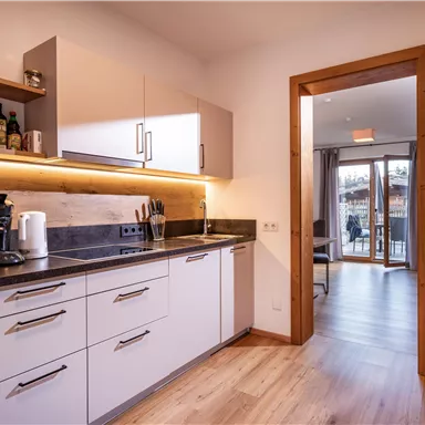 Eine moderne Küche mit einem Holzfußboden und einer hellen Beleuchtung. Durch die offene Tür ist ein Wohnbereich mit Zugang zum Balkon sichtbar.