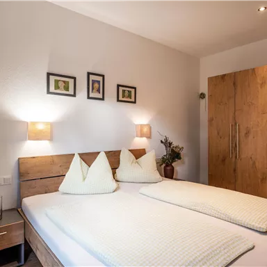 Ein gemütliches Schlafzimmer mit einem Doppelbett und sanften Beleuchtungen. An der Wand hängen einige Bilder, und ein Kleiderschrank steht im Raum.