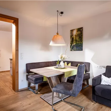 Ein gemütlicher Wohnbereich mit einem Esstisch, Stühlen und einer Sofaecke. An der Wand hängt ein Bild, und das Licht ist angenehm und warm.