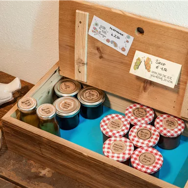 Eine Holzkiste mit verschiedenen Gläsern und Marmelade mit karierten Deckeln. Darin befinden sich auch kleine Schilder mit Preisen.