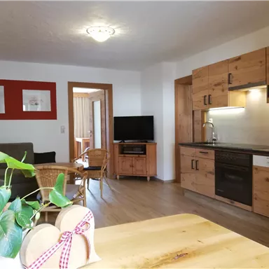 Eine moderne Küche mit Holzmöbeln und einem Esstisch. Im Hintergrund befinden sich ein gemütlicher Wohnbereich und ein Fernseher.