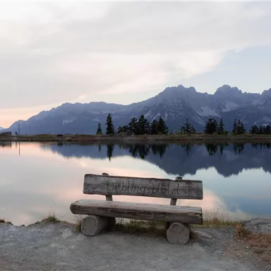 A wooden bench stands by the shore of a tranquil lake, surrounded by mountains. The sky has gentle colors at sunset.