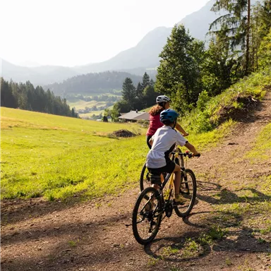 Two children are riding bicycles on a narrow path in a picturesque landscape. In the background, mountains and forests can be seen.