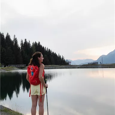 A woman stands at the shore of a tranquil lake and gazes into the distance. In the background, green forests and mountains can be seen.
