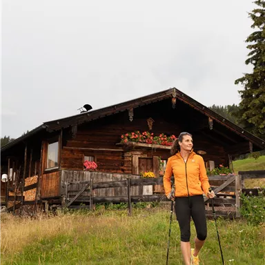 A woman in an orange jacket is walking in a meadow. In the background, there is a wooden house with flower boxes.