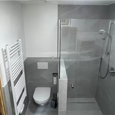 A modern bathroom with a glass shower and gray tiles. On the right, there is a toilet and a towel warmer.