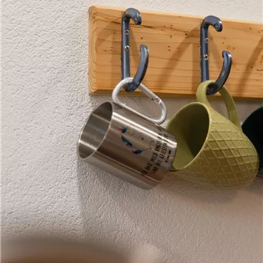 A wooden board with two hooks, from which a silver and a green cup are hanging. The cups have different designs and are placed on a white wall.
