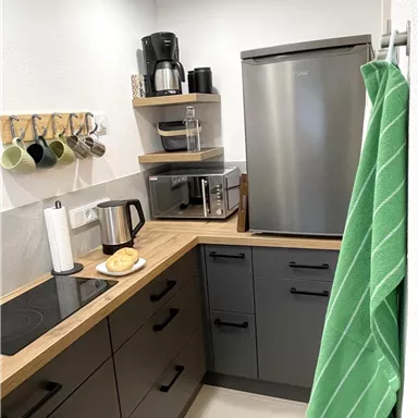 A modern kitchen with a wooden countertop, a refrigerator, and a microwave. Cooking utensils and mugs are neatly hung on the wall.