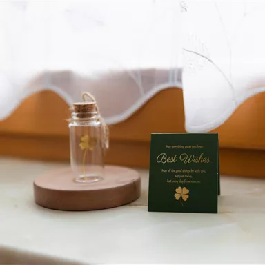 A glass with a golden four-leaf clover stands on a wooden coaster. Next to it lies a card with the inscription "Best Wishes".