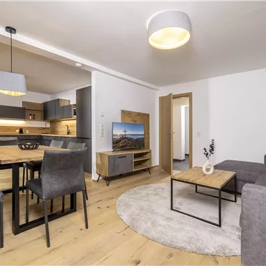 A modern living room with a cozy sofa and a wooden table. The open kitchen is well-equipped and the floor is made of light wood.