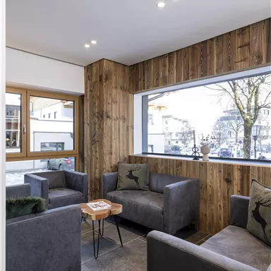 A modern living room with cozy gray sofas and a small wooden table. Large windows offer a view of an urban environment outside.