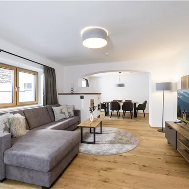 A modern living room with a gray sofa and a wooden table. In the background, there is a dining table and a TV unit.