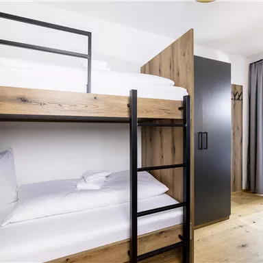 A modern room with a wooden bunk bed and a desk. Large windows provide plenty of daylight.