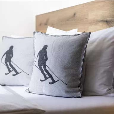 Two pillows with skier motifs are lying on a bed. The background is simple with wooden elements.
