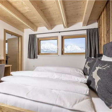 A cozy bedroom with wooden design and two windows that offer a view of the mountains. The beds are equipped with white sheets and decorative pillows.