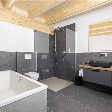A modern bathroom with a bathtub and a glass shower. The walls are in dark stone design and wooden ceilings give a warm atmosphere.