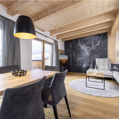 A modern living room with a wooden beam ceiling and large window. The room has a cozy seating area and a wooden dining area.