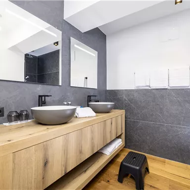 A modern bathroom with a wooden washbasin and two sinks. The walls are designed with gray tiles and there are large mirrors.