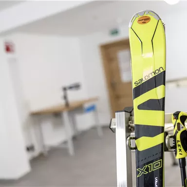 A pair of yellow skis with brand lettering is standing in a room. In the background, some furniture and a door can be seen.