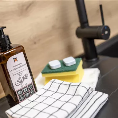 A bottle of dish soap stands next to a sponge and a cloth on a kitchen counter. The faucet in the background complements the clean kitchen ambiance.