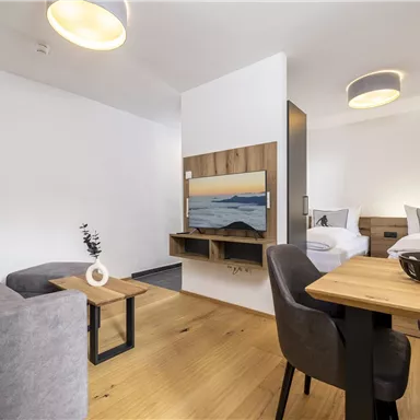A modern room with a cozy seating area and a dining table. A television is mounted on the wall, and large windows provide plenty of light.