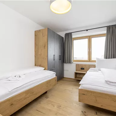 A bright room with two beds and a large window. The furniture is made of wood and the floor is carpeted.