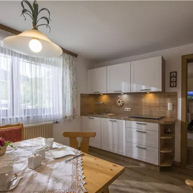 Eine helle, moderne Küche mit weißen Schränken und einem Esstisch. Das Fenster lässt viel Tageslicht herein und bietet einen Blick nach draußen.