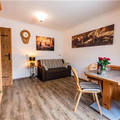 Ein gemütlicher Wohnraum mit Holzdekor, einem Sofa und einem Esstisch. An den Wänden hängen Bilder, und ein Blumenstrauß steht auf dem Tisch.