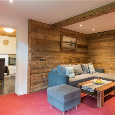 A cozy living room with wooden paneling and a red carpet. In the background, a dining area with a table and chairs is visible.