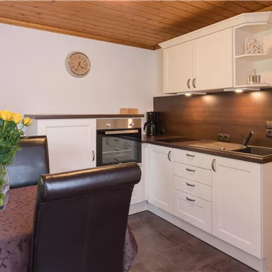 A modern kitchen with white cabinets and a dark countertop. On the table, there is a vase with fresh flowers and a clock on the wall.